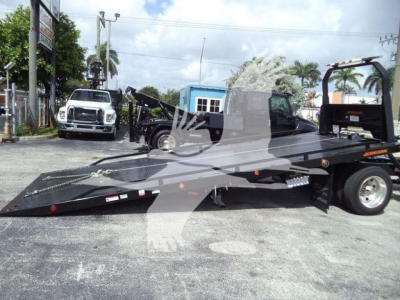 Flatbed tow truck trailer in a yard with another white truck in background; "MV" and equipment markings are visible on trailer.
