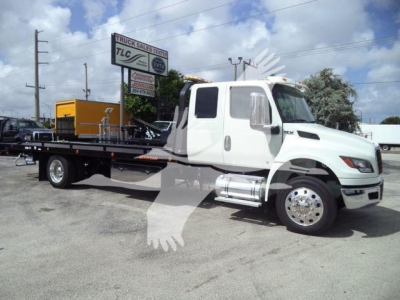 Heavy-duty white tow truck with flatbed in a dealership lot, International MV cab, chrome fuel tanks, large front grille, side mirrors, and black flatbed with metal supports.