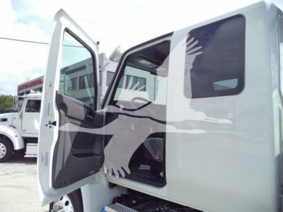 Truck cab interior visible; open door shows driver seat and steering area with side mirror. Background shows other trucks and dealership lot.