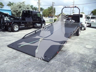 Equipment trailer bed with a tilted ramp and winch straps, black flatbed surface, white railing at rear, on a dealership lot.