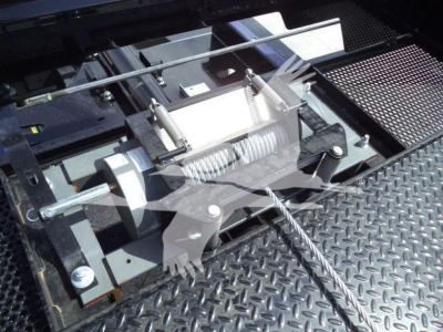 Trailer winch and mounting hardware with steel cable and threaded rods on a diamond plate surface. Visible components include a winch drum, spring-loaded tensioner, and mounting brackets.