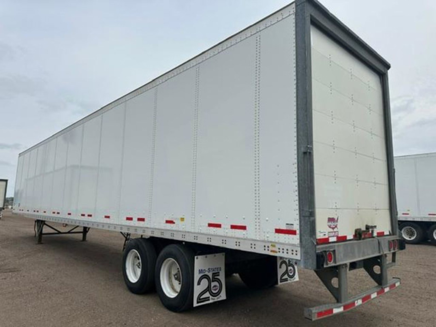 Trailer side view showing a white dry van trailer with tandem axles, riveted panels, and rear doors. Visible decals include '2 6' placards and small red/white reflective tape along the lower edge.