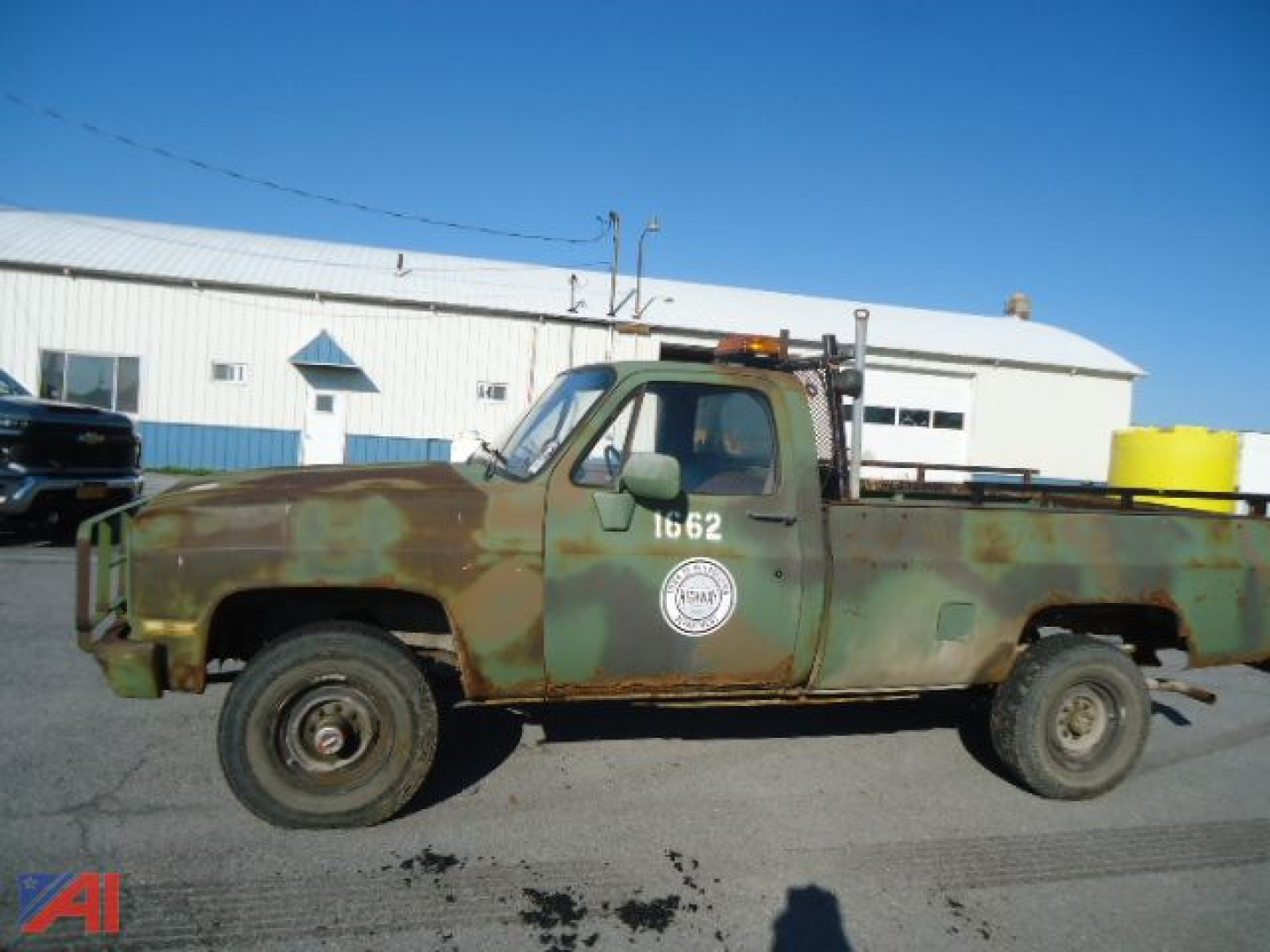 1985 Chevrolet military D30 pickup truck side view with weathered camouflage paint, white circle logo on door, light bar on cab, flatbed, in a lot.
