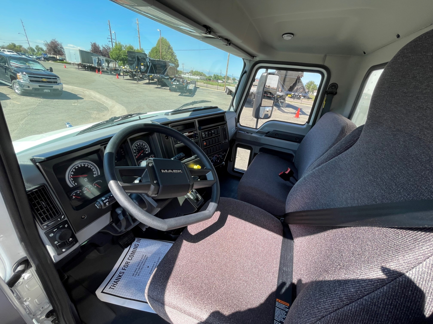 Cab interior of a 2025 Mack MD6 truck showing steering wheel, dashboard with gauges, and fabric seat. Side window and door panel visible; sunlight through windshield.