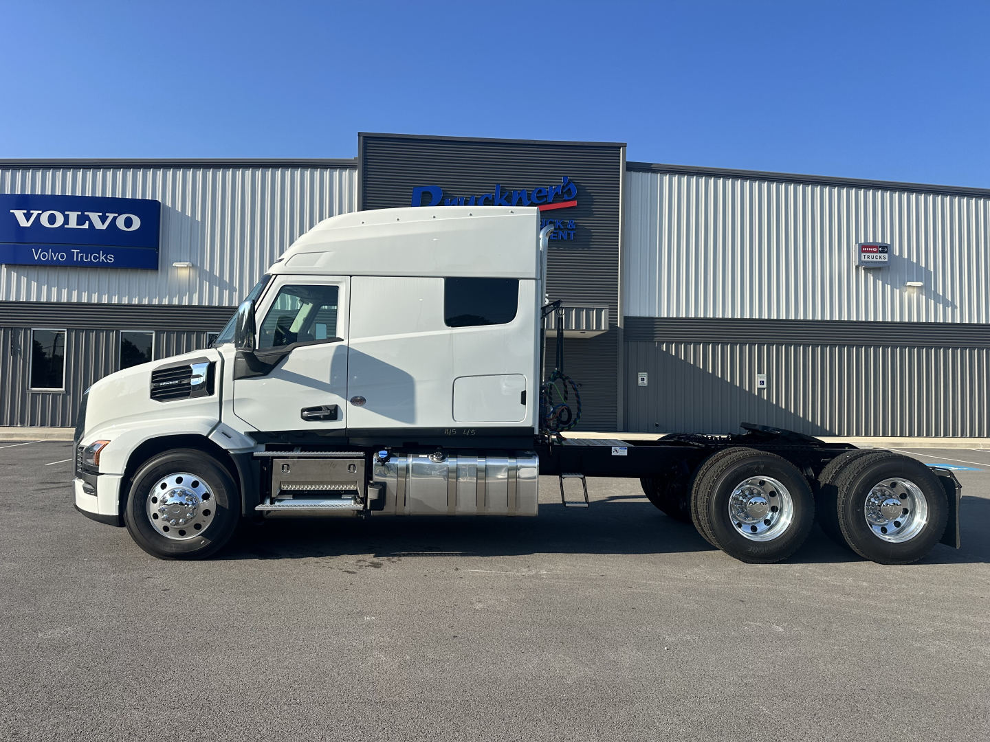 Truck cab side profile of a white 2027 Mack PR64T76M tractor with sleeper, aluminum fuel tanks, and six wheels visible, dealership building in background.