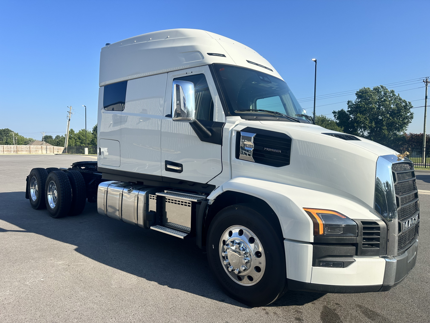 Truck cab side and front view showing a white 2027 Mack PR64T76M conventional sleeper with chrome wheels, fuel tanks, and dual rear axles. Visible badges and design details
