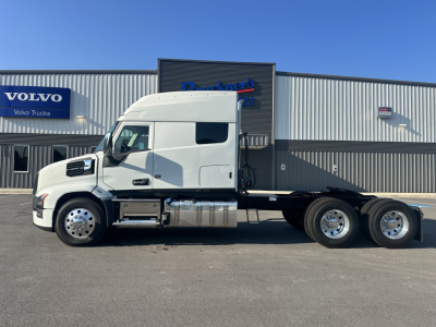 Truck cab side profile of a white 2027 Mack PR64T76M tractor with sleeper, aluminum fuel tanks, and six wheels visible, dealership building in background.