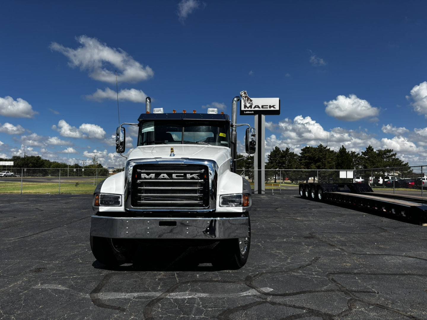 Mack truck front view with grille and Mack emblem; dealership sign in background; clear sky.