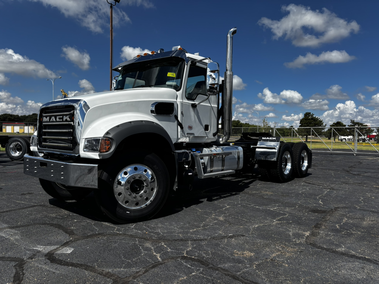 Truck: 2027 Mack GR64FT conventional daycab, white cab, chrome exhaust stacks, dual rear axles, front grille with Mack badge, visible tires, dealership lot.