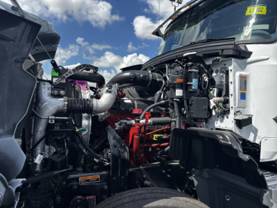 Front engine bay view of a new 2027 Mack GR64FT daycab truck showing engine components and open hood area.