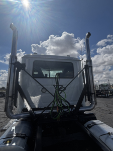 Truck cab of a 2027 Mack GR64FT in-view from behind with chrome vertical exhaust stacks and green hoses on the star-of-daycab, front window visible.