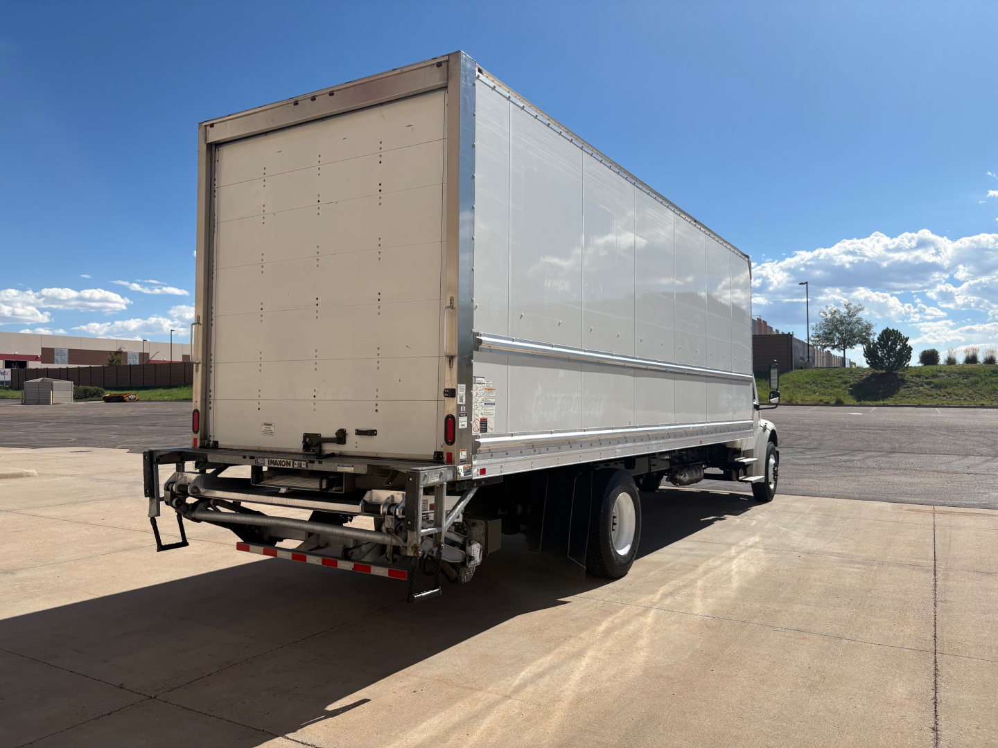 Freightliner M2-106 box truck with rear lift gate and silver cargo box, dual rear wheels, parked on concrete under blue sky.