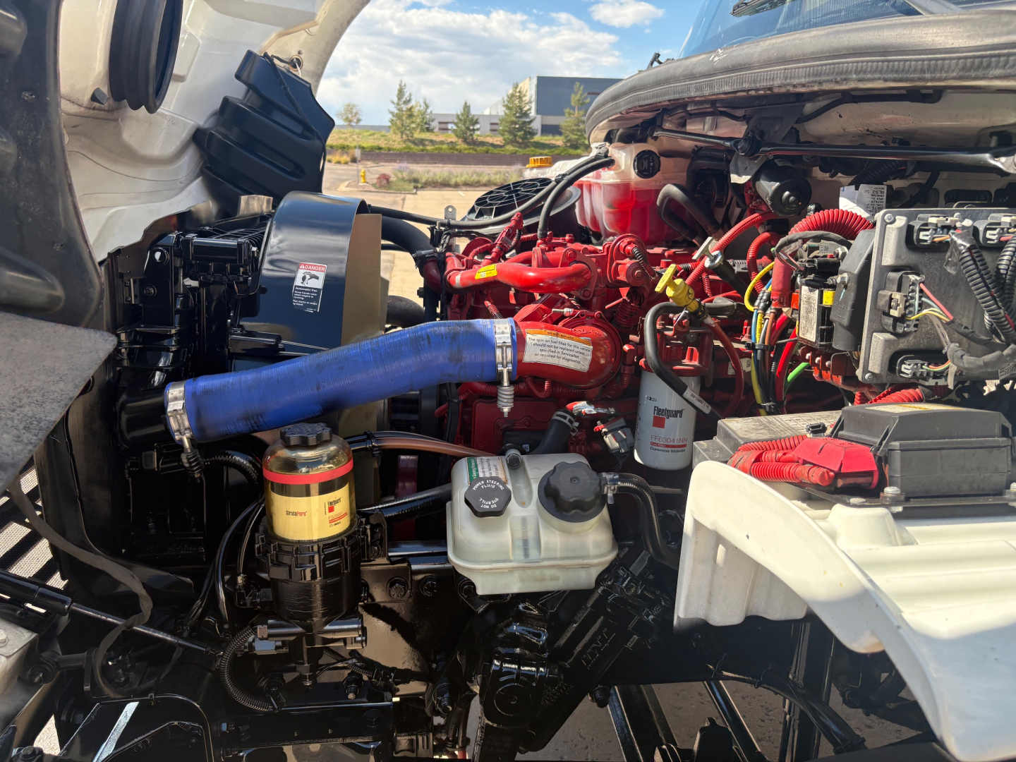 Freightliner M2-106 engine bay in-view showing red engine block, blue hose, and various fluid reservoirs and wiring harnesses.