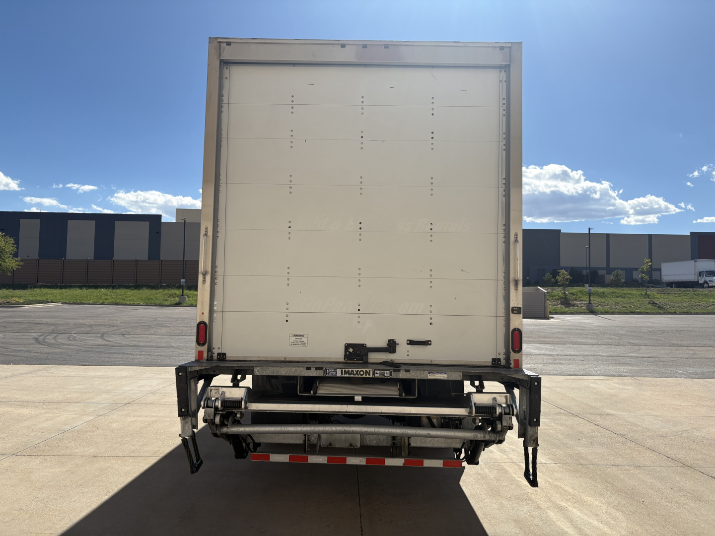 Rear view of a Freightliner M2-106 box truck showing a white cargo door, rear bumper, and metal undercarriage with reflective red/white tape.