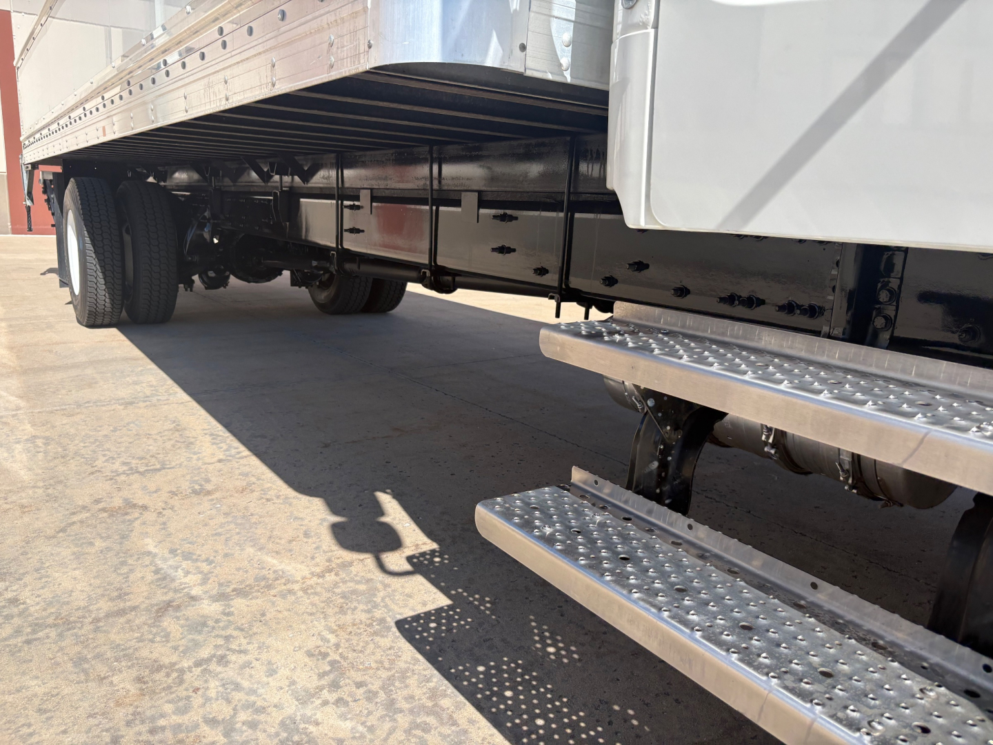 Rear/side view of a Freightliner M2-106 truck with aluminum side rails and metal step tread. Visible undercarriage includes dual rear wheels, suspension, and frame.