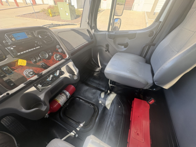 Freightliner M2-106 cab interior showing dashboard with controls, steering wheel, gray driver seat, and open floor area with a red tool box and extinguisher nearby.