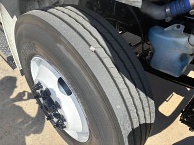Freightliner M2-106 front wheel and suspension area showing tire, lug nuts, and blue coolant reservoir in engine bay.