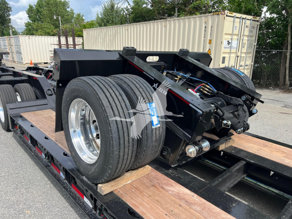 Trailer axle assembly on a flatbed lowboy trailer showing dual tires, hydraulic components, and steel frame with wooden deck sections.