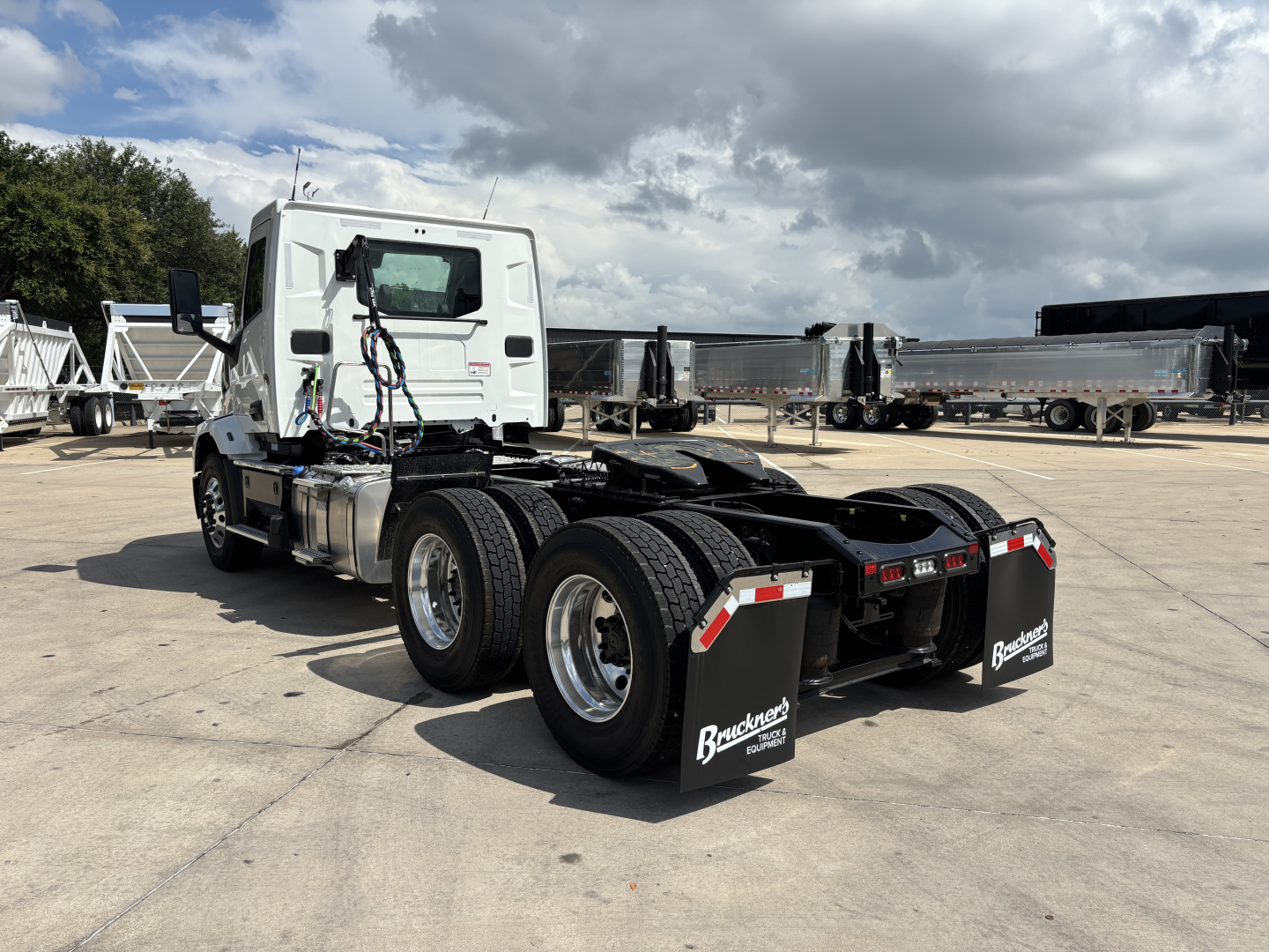 Truck chassis and cab of a white 2027 Mack AN64T tractor from rear-left perspective; dual rear axles with black mud flaps showing Bracken? branding; several trailers in background.