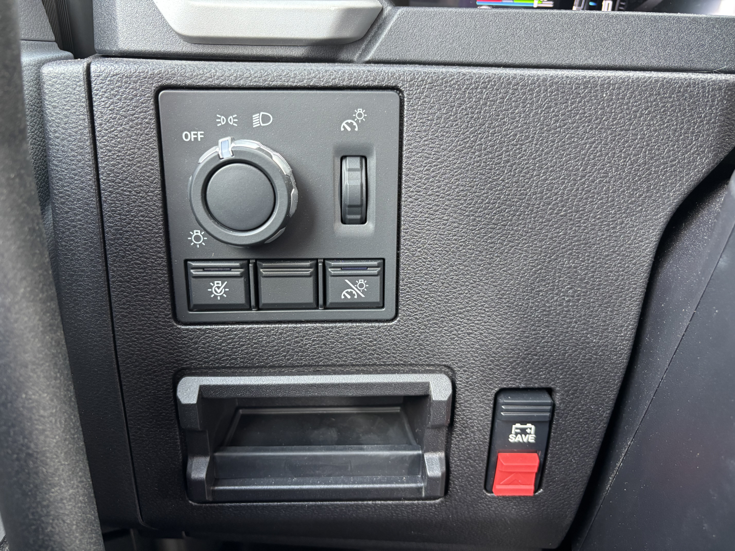 Dashboard control panel of a 2027 Mack AN64T showing headlight dial, rotary dimmer, and five switch buttons; small cubby and a red 'Save' button on the right.