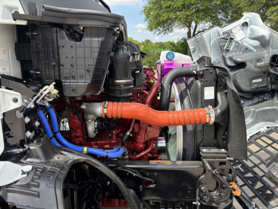 Engine bay of a 2027 Mack AN64T showing orange flexible coolant hose, blue hoses, red engine block, radiator, and surrounding components.