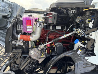 Truck engine bay showing pink coolant reservoir, pink washer fluid, orange air filter hose, red engine block, blue dipstick, and various hoses and components in a Mack AN64T engine bay.