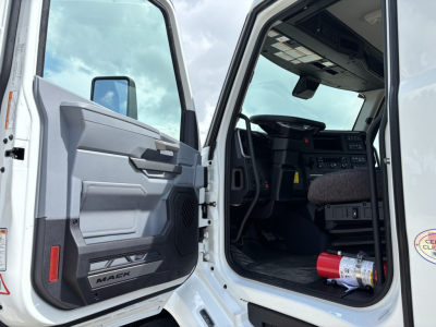 Truck cab interior and open door of a new Mack AN64T day cab showing dashboard, steering wheel, and front seats; visible label 'MACA' on door panel.