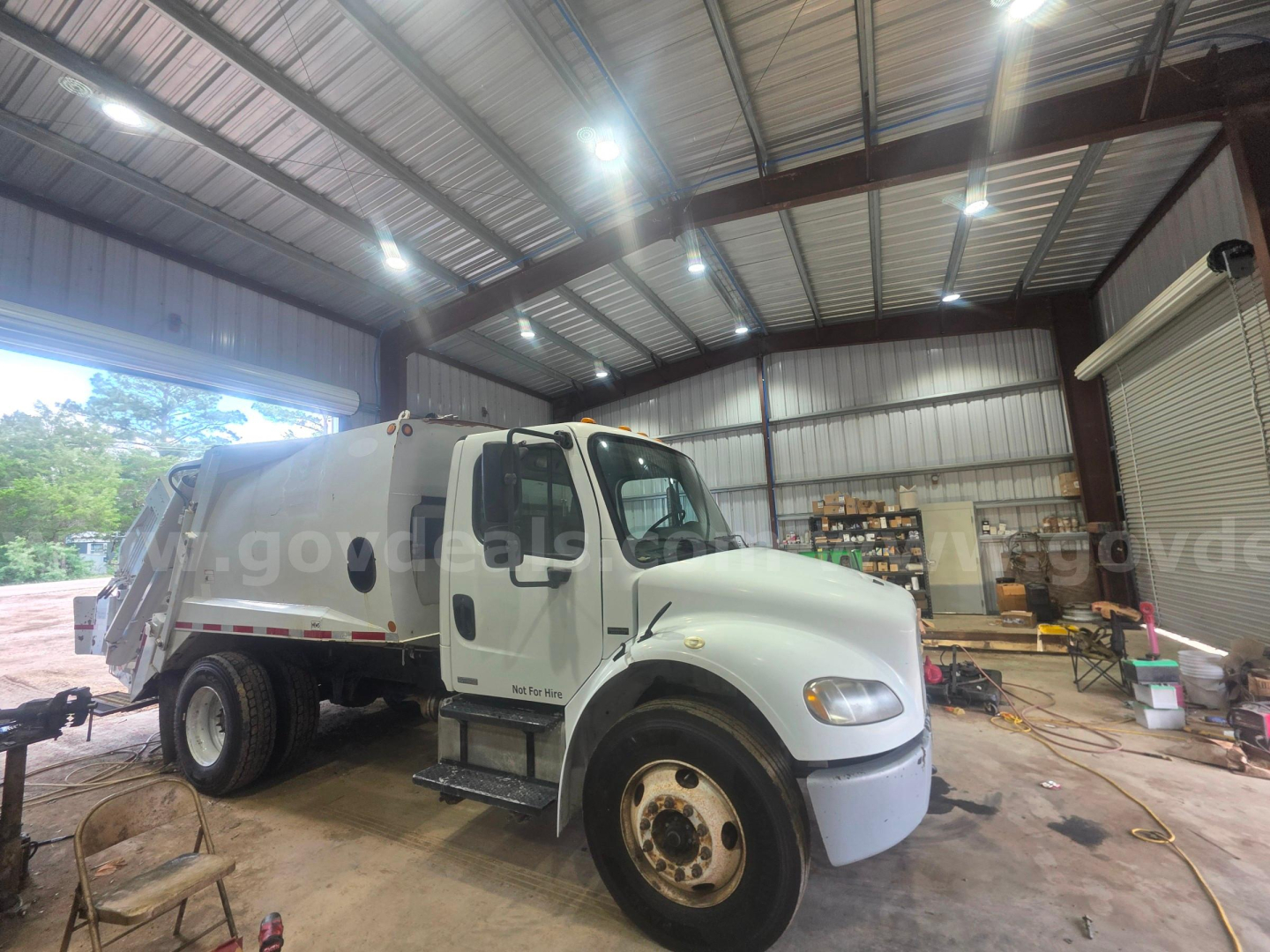 Front three-quarter view of a white Freightliner M2 106 garbage truck inside a warehouse, with side cab steps visible and storage shelves in background.