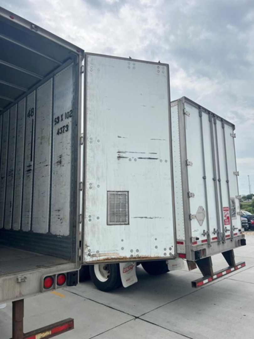 Trailer van rear view showing two rear doors of an enclosed cargo trailer and open trailer on left; weathered metal surfaces and reflective safety decals present.