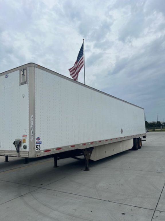 Utility van trailer showing long white box with riveted metal sides, rear doors, support legs, and an American flag in the background.