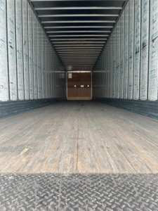Trailer interior showing empty cargo area with wooden floor and metal sidewalls, rear ramp opening visible.