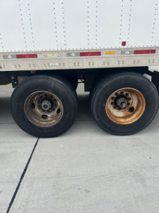Trailer dual rear axles with wheels; rusty wheel on right and left with black tires, silver rim on left, white box trailer with dot rivets above.