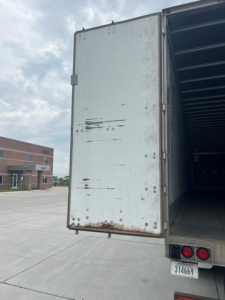 Rear of a utility van trailer showing the open cargo area and rear doors with some rust and wear. Visible text includes the trailer rear lights and license plate area.