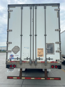 Rear view of a white utility van trailer with double doors, hinges, latches, and warning label; bumper and taillights visible.