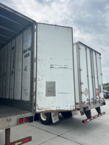 Trailer van rear view showing two rear doors of an enclosed cargo trailer and open trailer on left; weathered metal surfaces and reflective safety decals present.