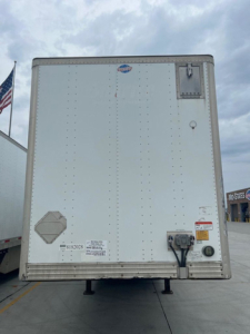 Back view of a white box truck trailer showing rear doors with latching hardware and a small circular vent, various stickers, and a hitch area.