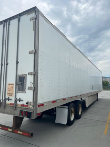 Semi-trailer box trailer in service showing plain white exterior and rear doors with CAUTION label and taillights; dual rear wheels visible under the trailer.
