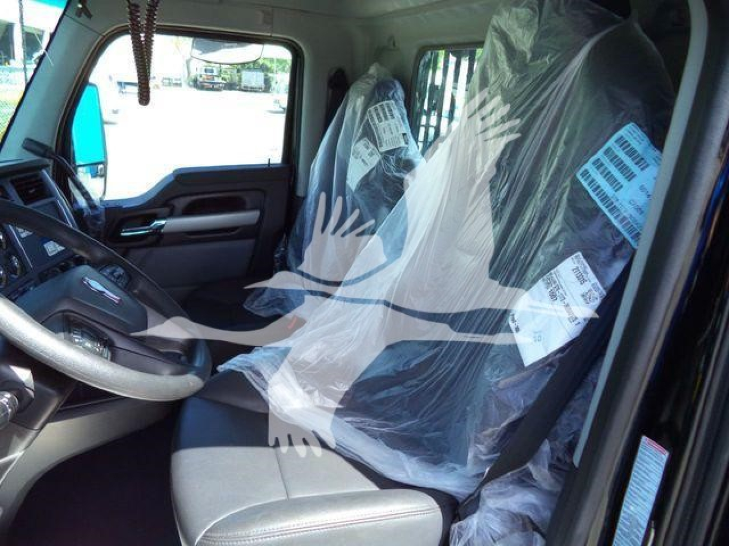 Truck cab interior showing driver seat and passenger seat covered in plastic. Steering wheel visible with Kenworth branding; plastic wrapping on seats and some tags hanging from the cabin.