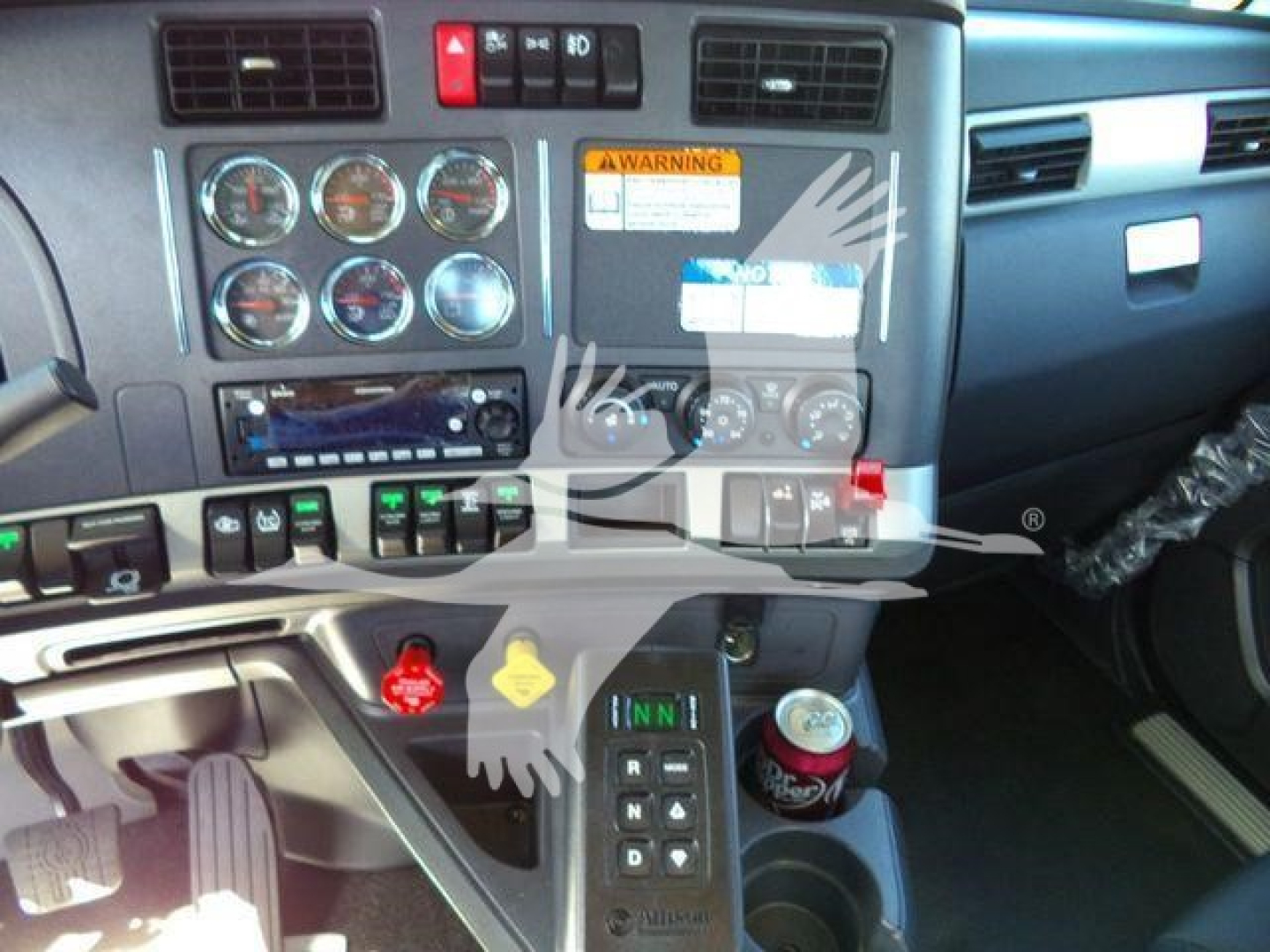 Dashboard area of a 2026 Kenworth T880 showing gauges, warning labels, control buttons, beverage can in cup holder, and gear/section with switches.