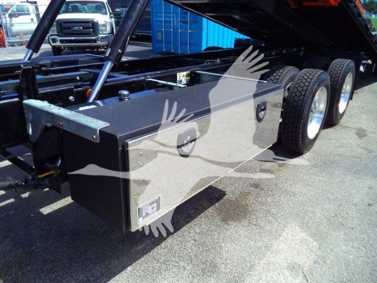 Flatbed truck undercarriage with a metal toolbox mounted near the rear wheels; multiple wheels visible; hydraulic components and frame structure; some small labels on toolbox.