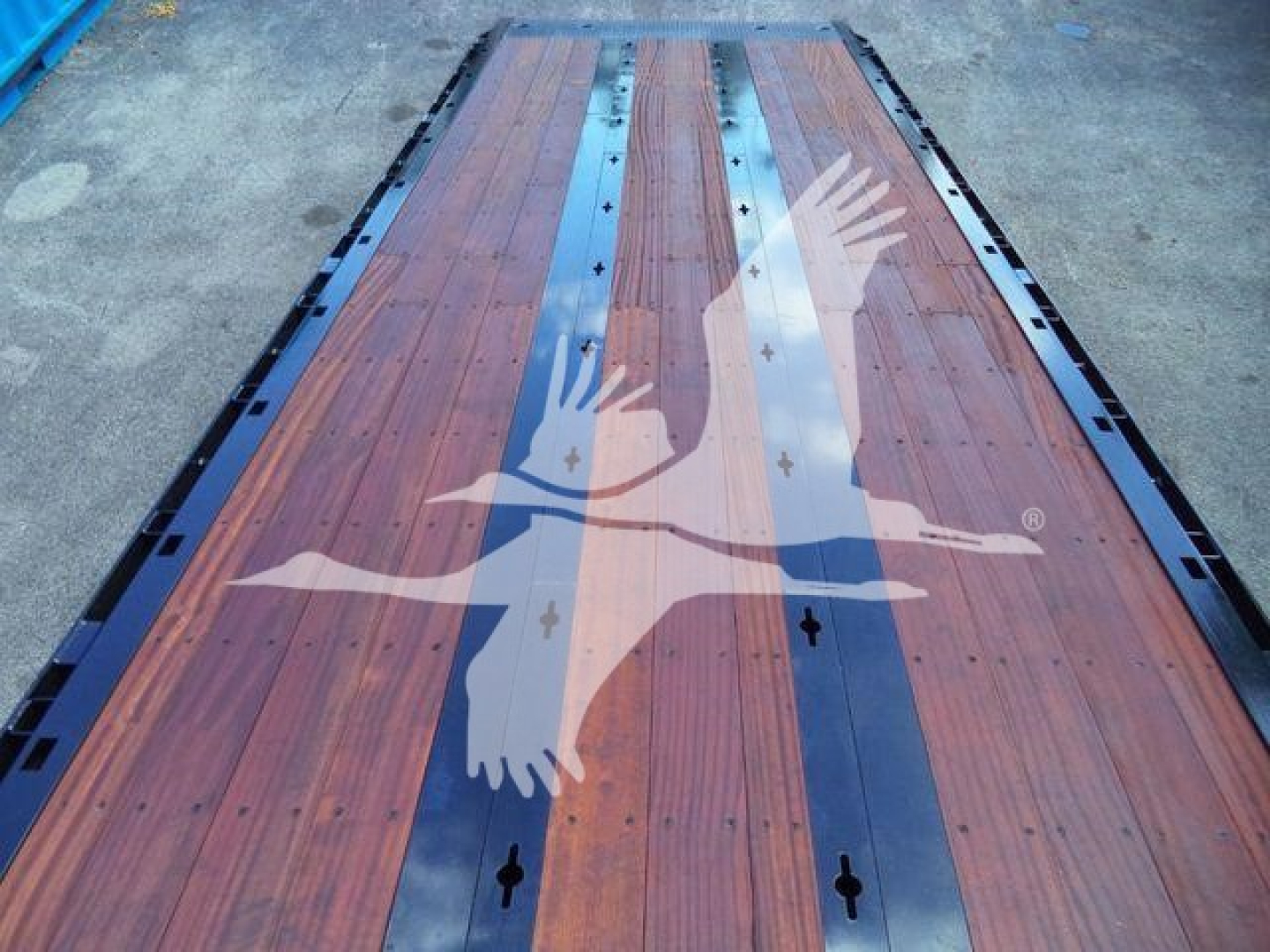 Flatbed trailer or loading deck with wooden planks and blue steel side rails showing a large translucent logo of a flying eagle or angel figure overlaying the surface.
