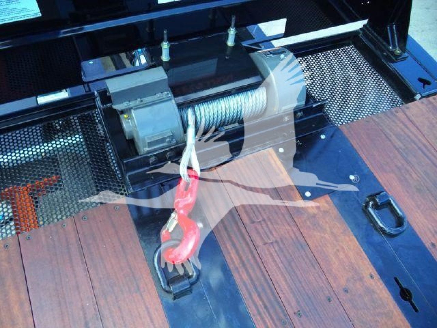 Winch assembly mounted on truck bed deck with hook and winch cable. Visible components include a motor, drum, and steel roller equipment on a blue-metal surface.