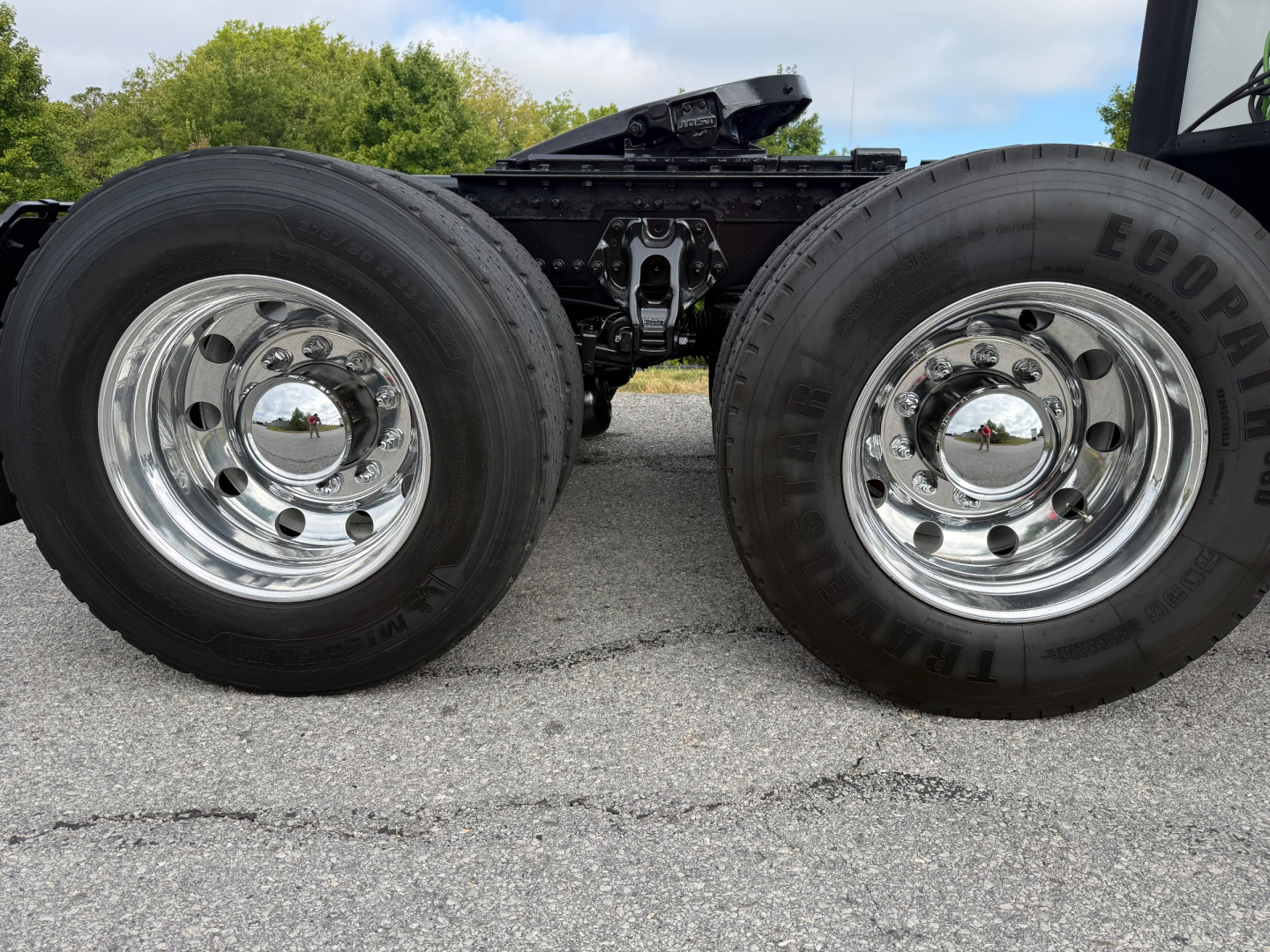 Dual rear wheels on a Kenworth truck chassis showing chrome hubcaps and tires; part of the drivetrain and suspension visible between wheels.