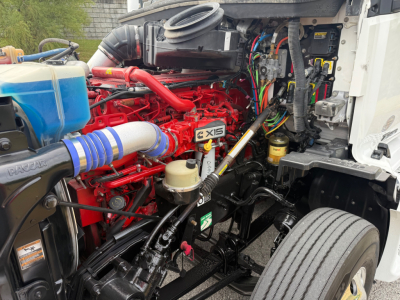 Kenworth T680 engine bay showing red engine block with X15 badge, blue coolant reservoir, black chassis, and various hoses and wiring harnesses.