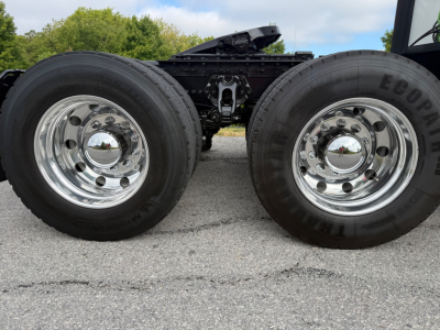 Dual rear wheels on a Kenworth truck chassis showing chrome hubcaps and tires; part of the drivetrain and suspension visible between wheels.