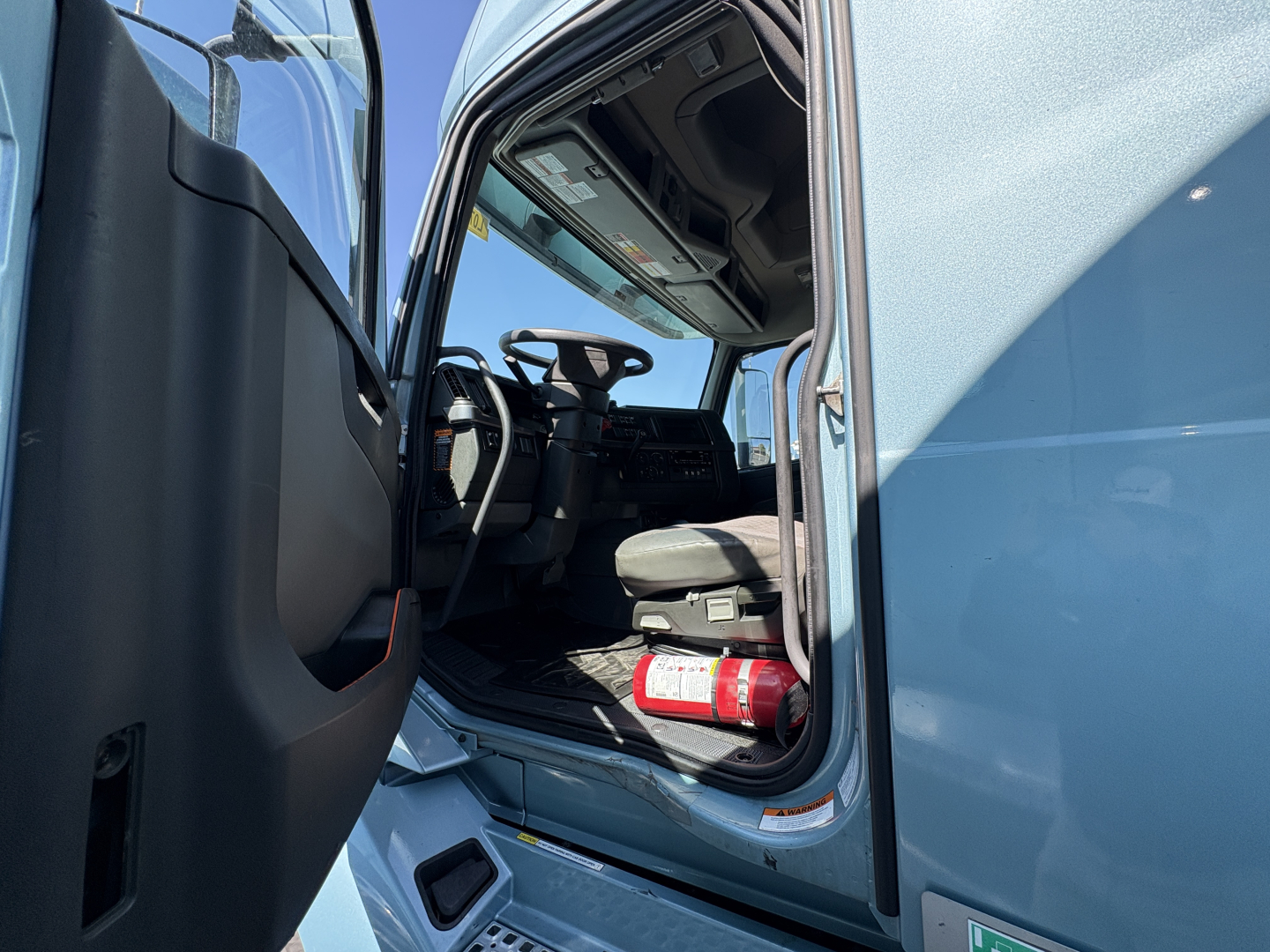 Truck cab interior of a 2022 Volvo VNL64T760 from the driver’s side door open, showing steering wheel, driver seat, dashboard, and a fire extinguisher near the entry.