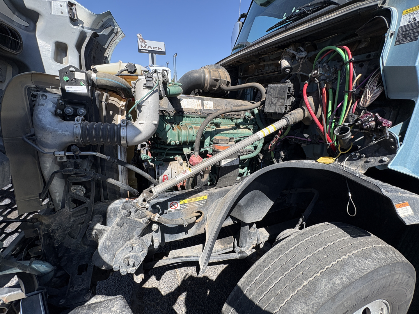 Volvo VNL64T760 engine bay showing multiple colored wires, hoses, and components under the hood; view includes front wheel and exposed mechanicals.
