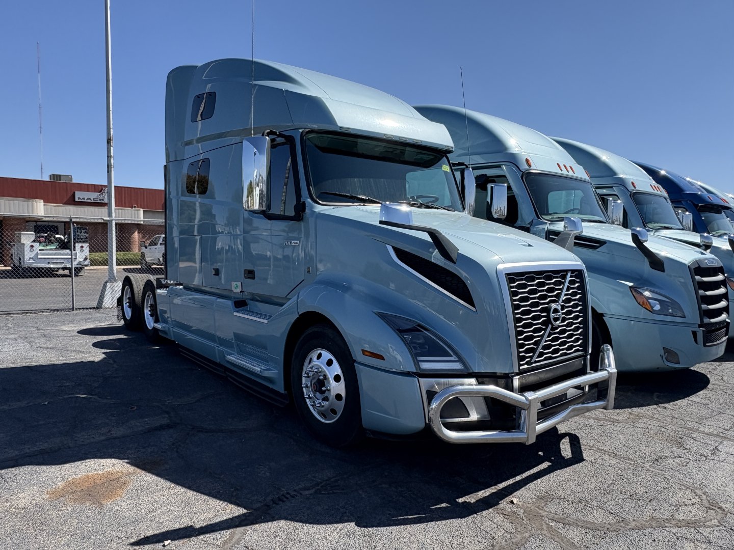 Truck front view of a blue 2022 Volvo VNL64T760 sleeper cab showing chrome grille guard and nearby similar trucks.