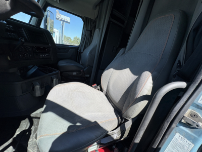 Truck cab interior of a 2022 Volvo VNL64T760 showing driver seat and passenger seat with grey upholstery, dashboard controls, open door frame, and sunlight through window