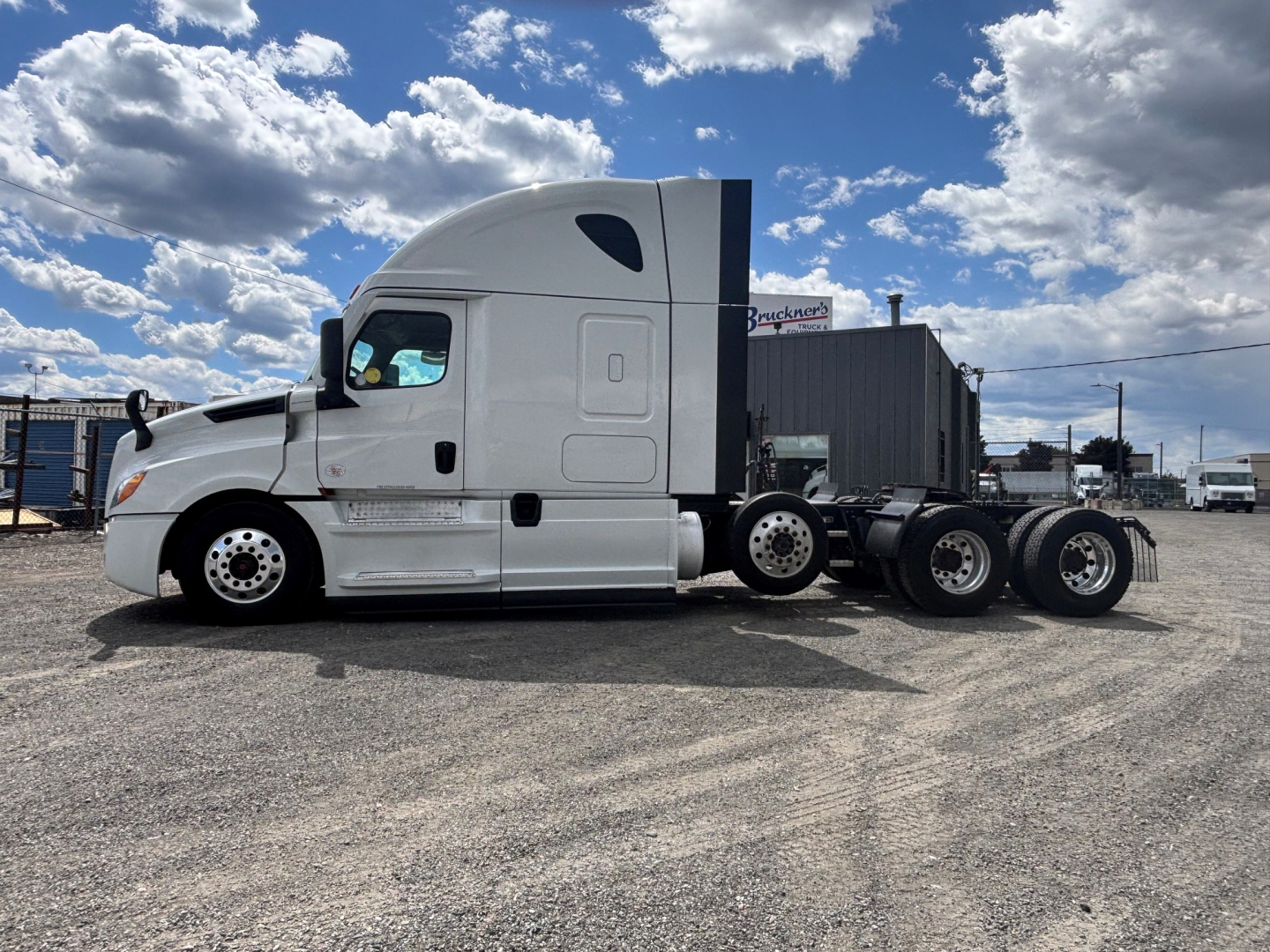 Truck side view showing a white Freightliner CA-126 tractor with sleeper cab and multiple rear axles on a gravel lot under blue sky.